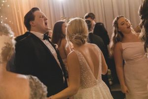 Joyful wedding guests singing and dancing with bride in pearl-embellished gown at reception.