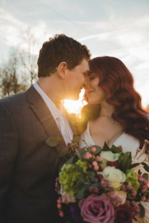 Romantic bride and groom nearly kissing at golden sunset with a colorful floral bouquet in the foreground.