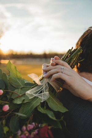 Close-up of bride's hands holding a green wedding bouquet with engagement and wedding rings at golden hour.