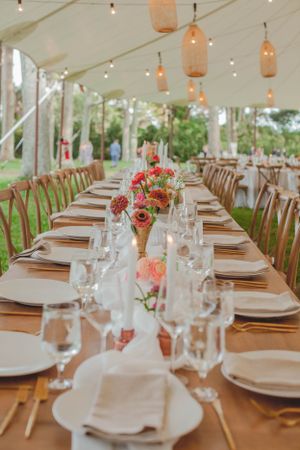 Elegant outdoor wedding reception tent with long tables, gold flatware, floral centerpieces, and string lights.