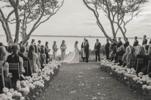 Outdoor wedding ceremony aisle lined with white roses leading to couple at altar near waterfront.