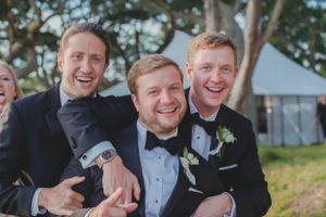 Three groomsmen in tuxedos laughing and embracing outdoors at wedding reception.