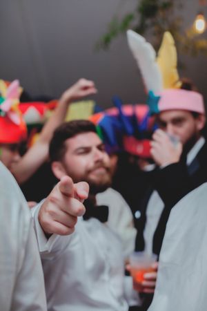 Bearded groomsman in white shirt and bow tie pointing at camera during festive wedding reception party.