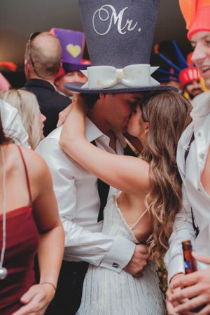 Couple sharing a kiss at a lively wedding reception with guests wearing fun party hats and props.