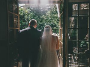 Bride and father standing at open doors overlooking lush green trees before wedding ceremony.