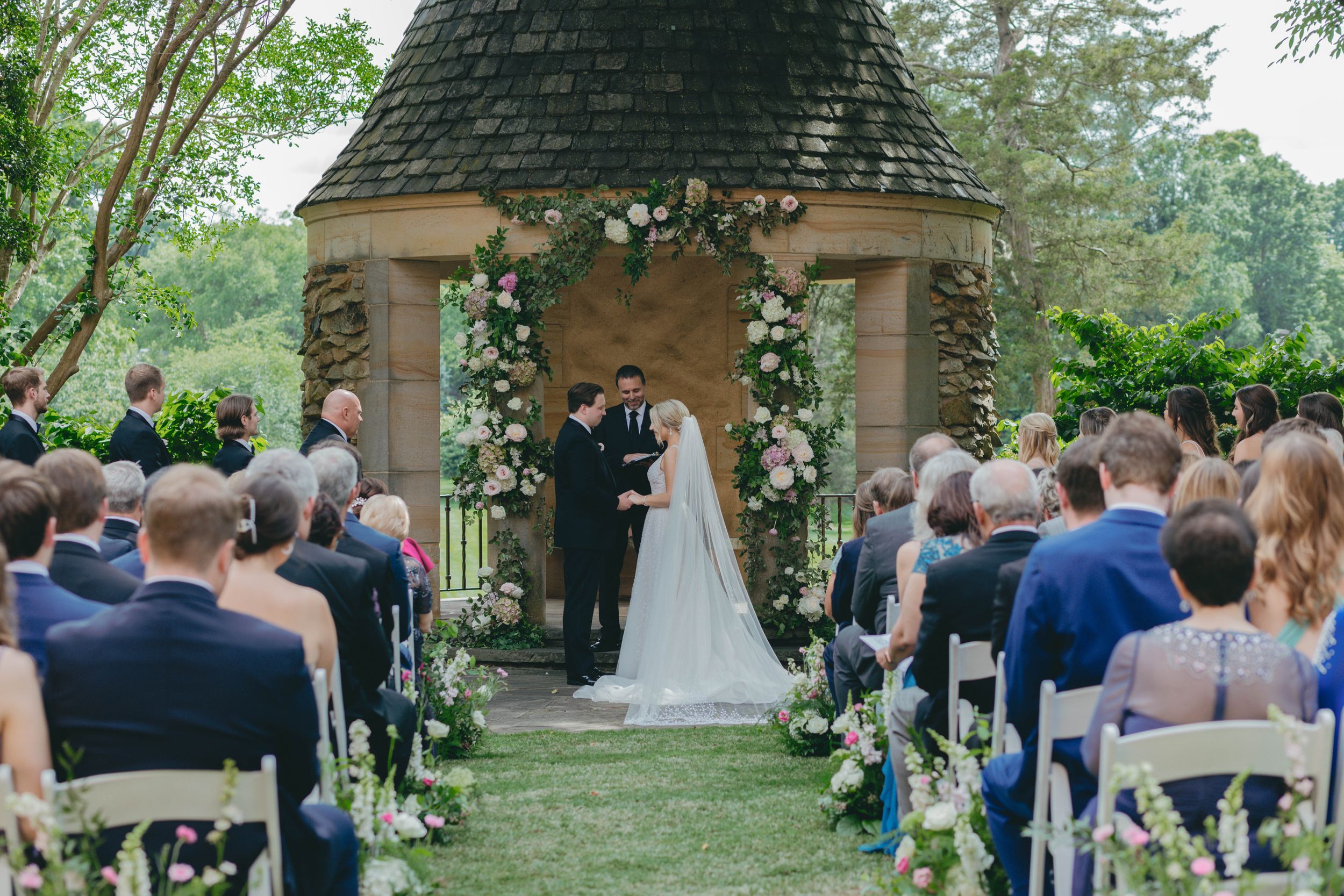 Outdoor wedding ceremony at stone chapel with floral arch, guests seated in garden setting.