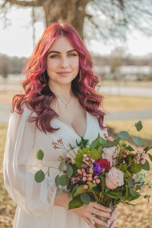 Bride with vibrant red wavy hair holding a colorful wedding bouquet of roses and greenery in a white dress outdoors.