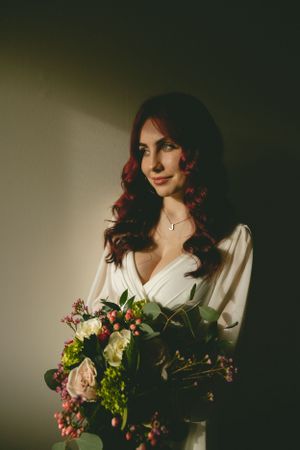 Elegant bride with red hair in a white dress holding a lush floral bouquet in dramatic moody indoor lighting.