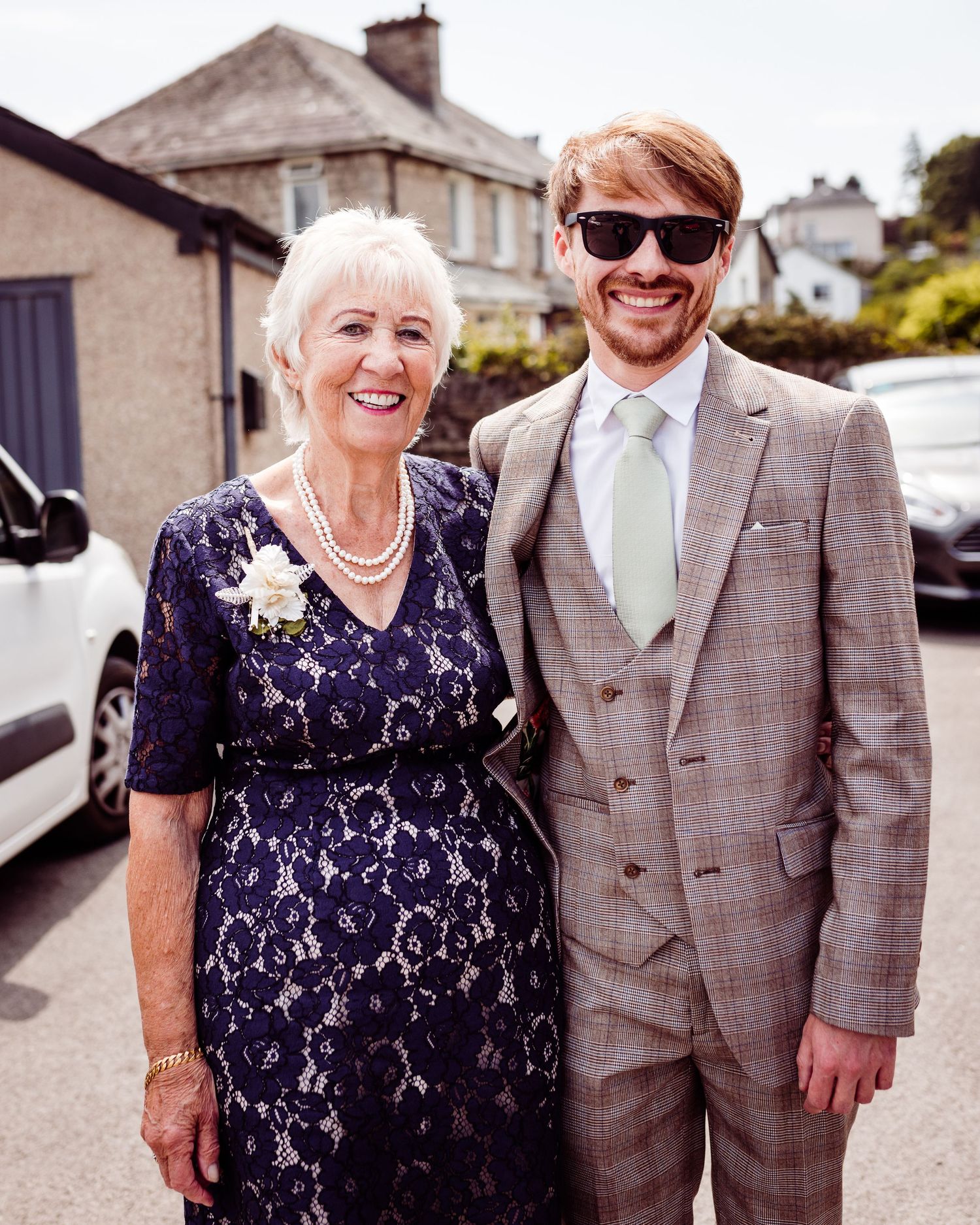 Elegant wedding attire featuring navy lace dress and light grey suit at outdoor venue.