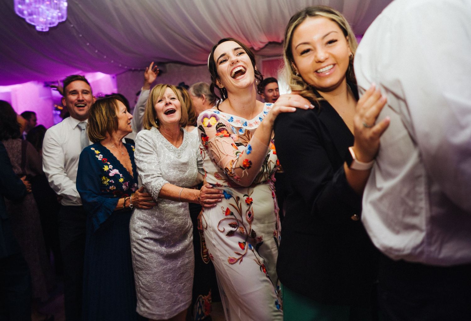Lively reception scene with guests dancing and laughing under purple lighting at a wedding celebration.