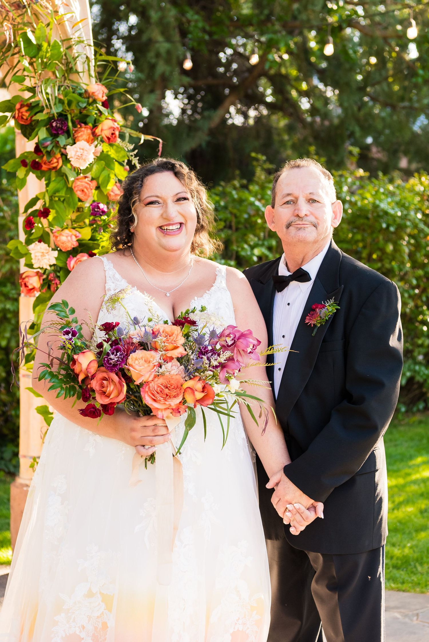 A bride in a lace dress holding the hand of her father during family formal photos at her outdoor wedding.