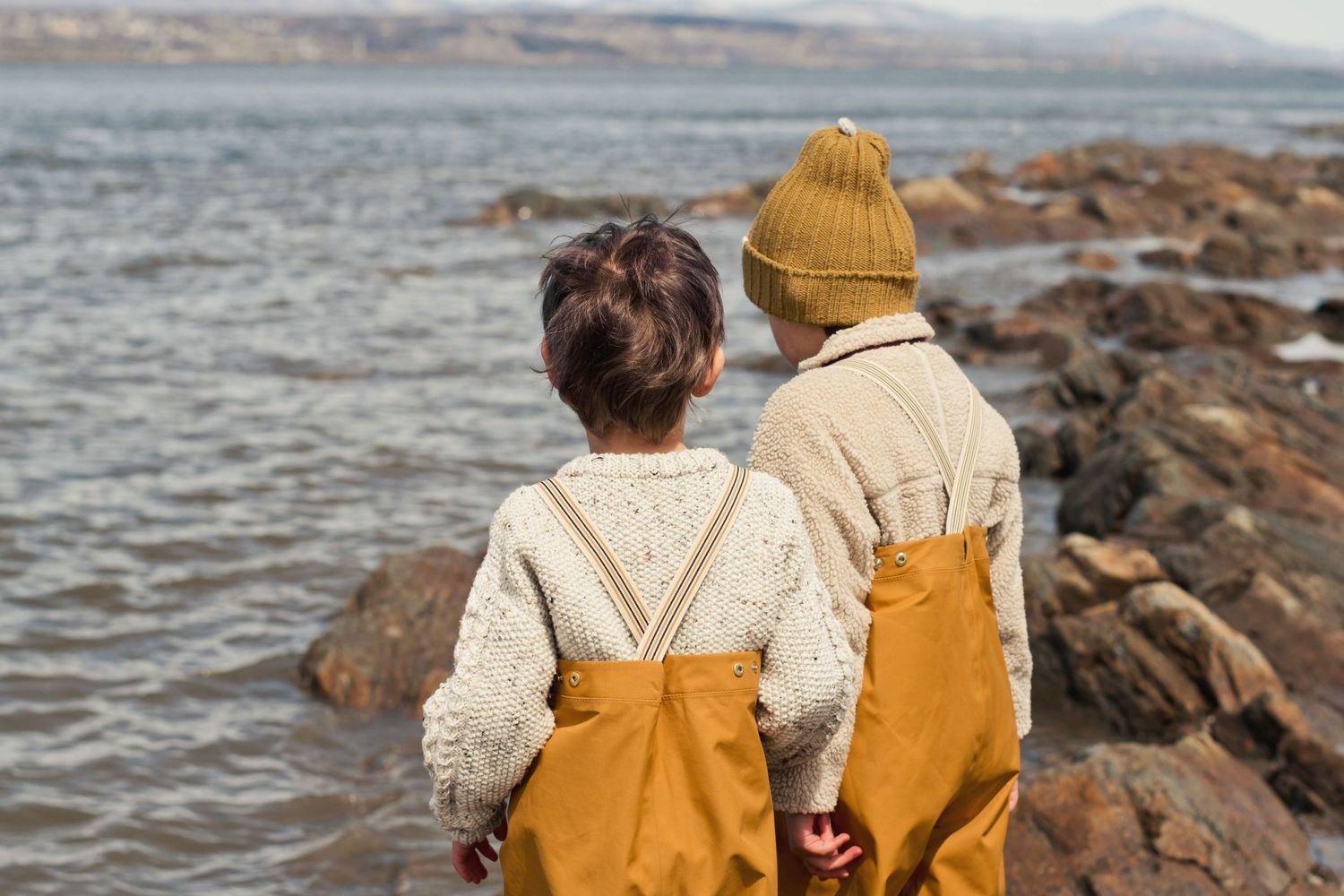 Two figures in matching cream sweaters and yellow overalls looking at ocean view.