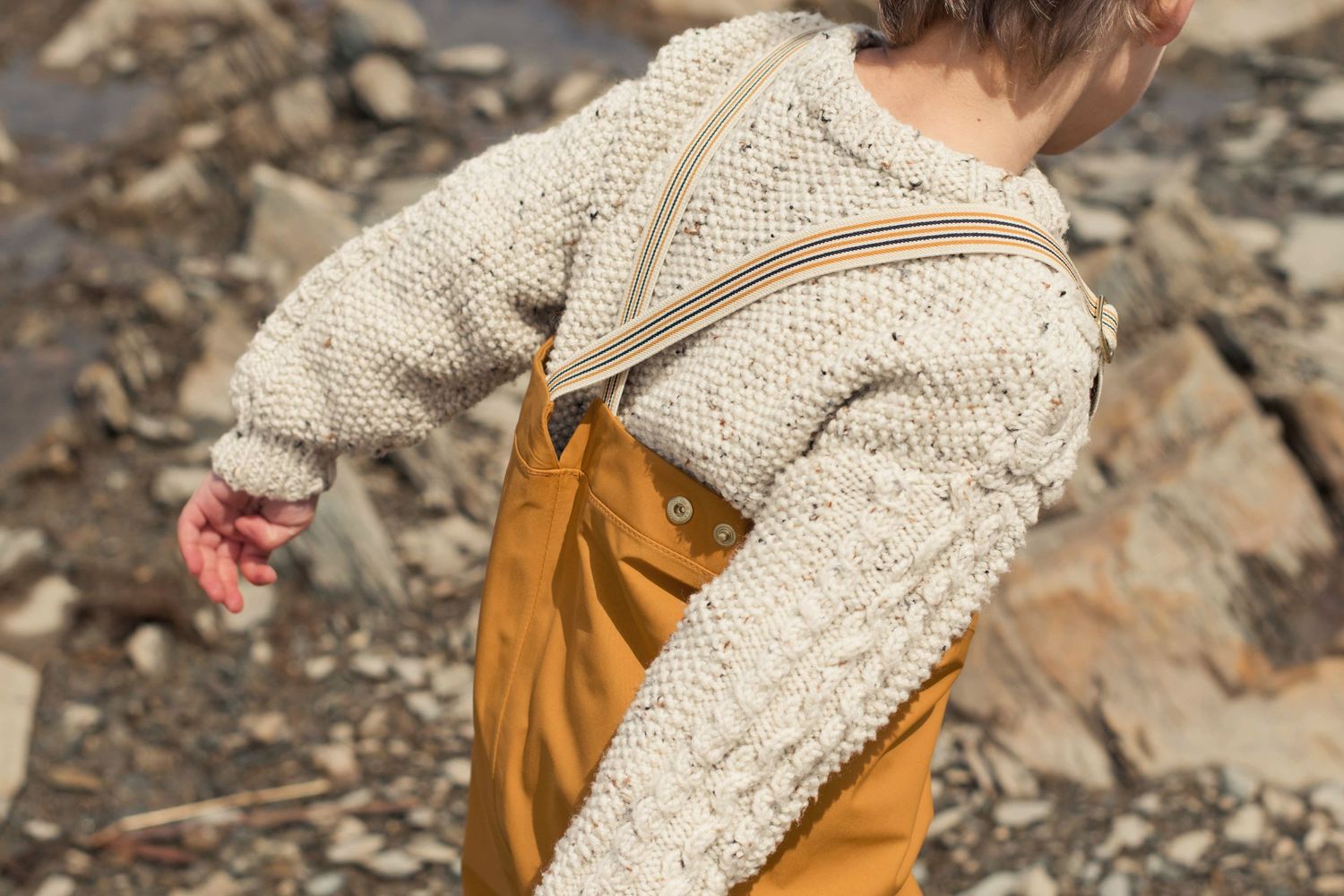 Close-up of knit sweater and orange overalls against rocky beach background.