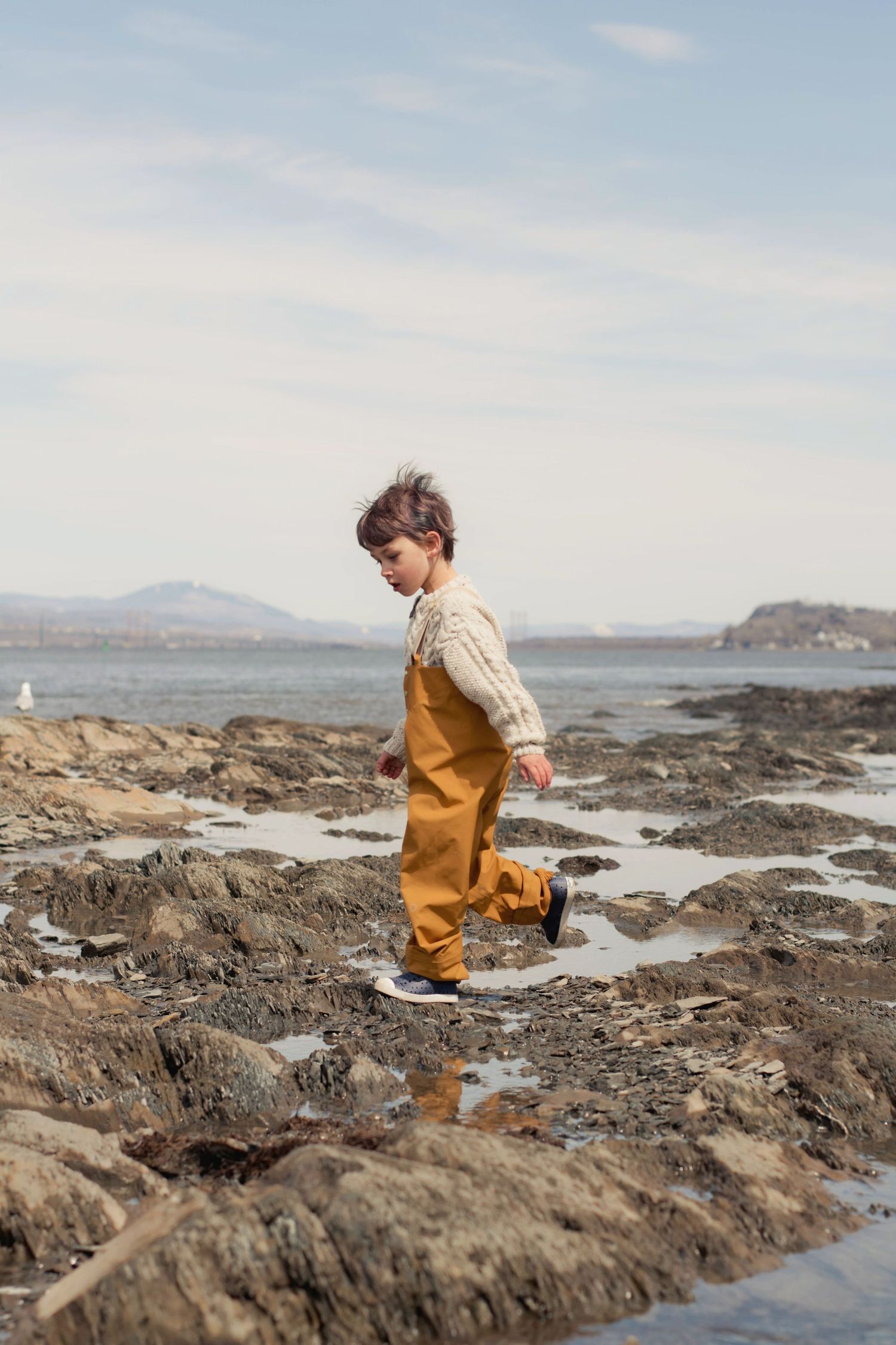 Young explorer in orange overalls standing on rocky coastal shoreline.