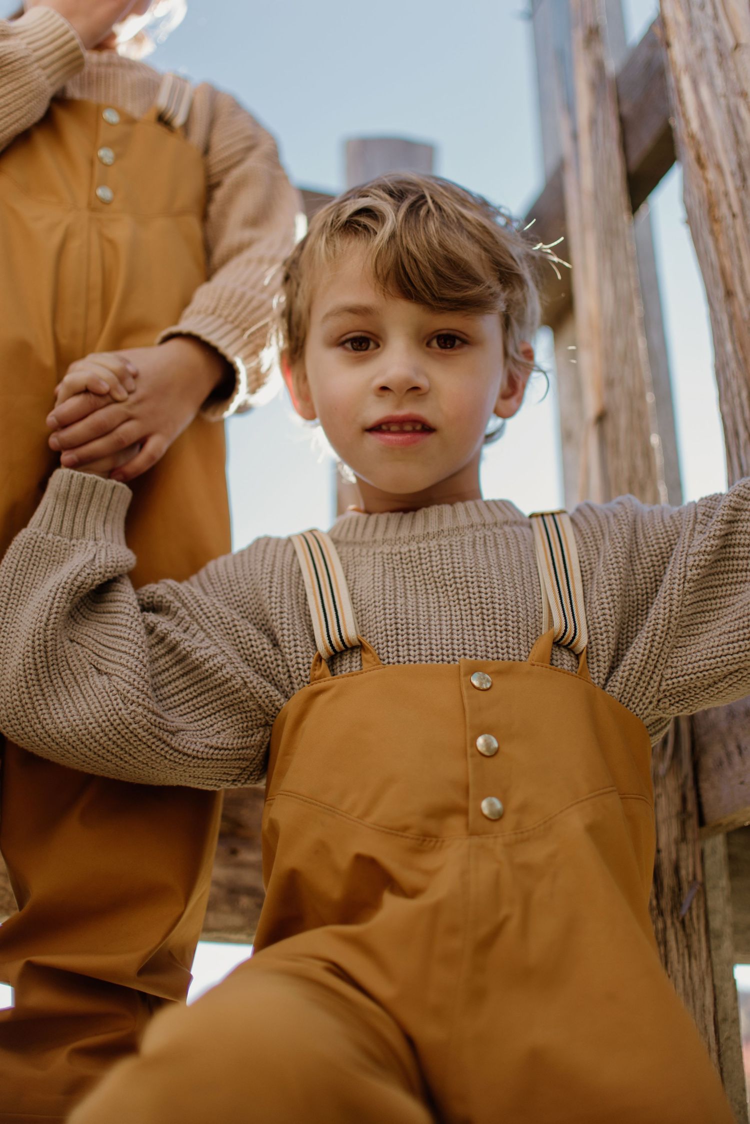 Child in beige sweater and brown overalls poses against wooden structure outdoors.