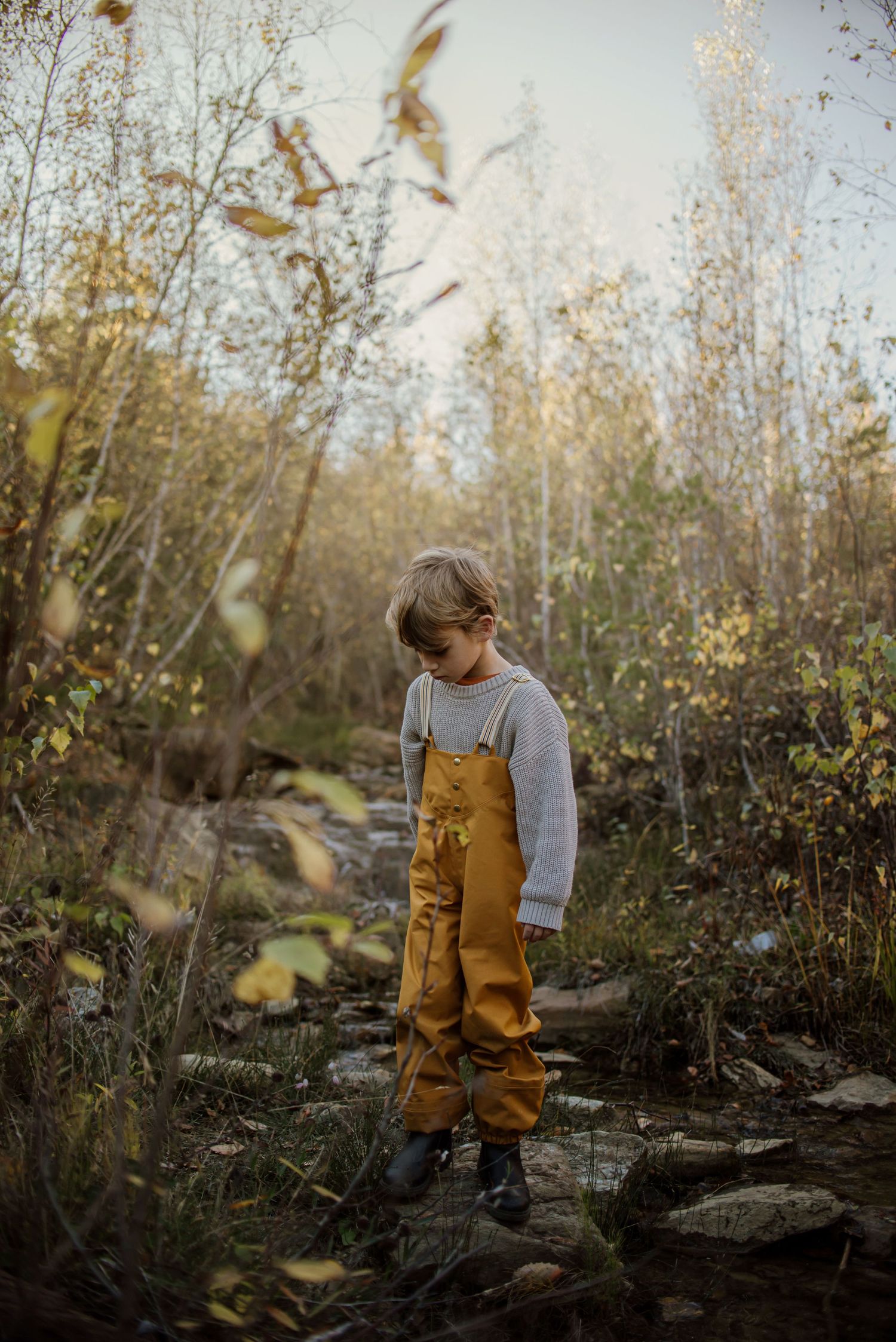 Child in gray sweater and mustard overalls standing on rocky forest path amid fall foliage.