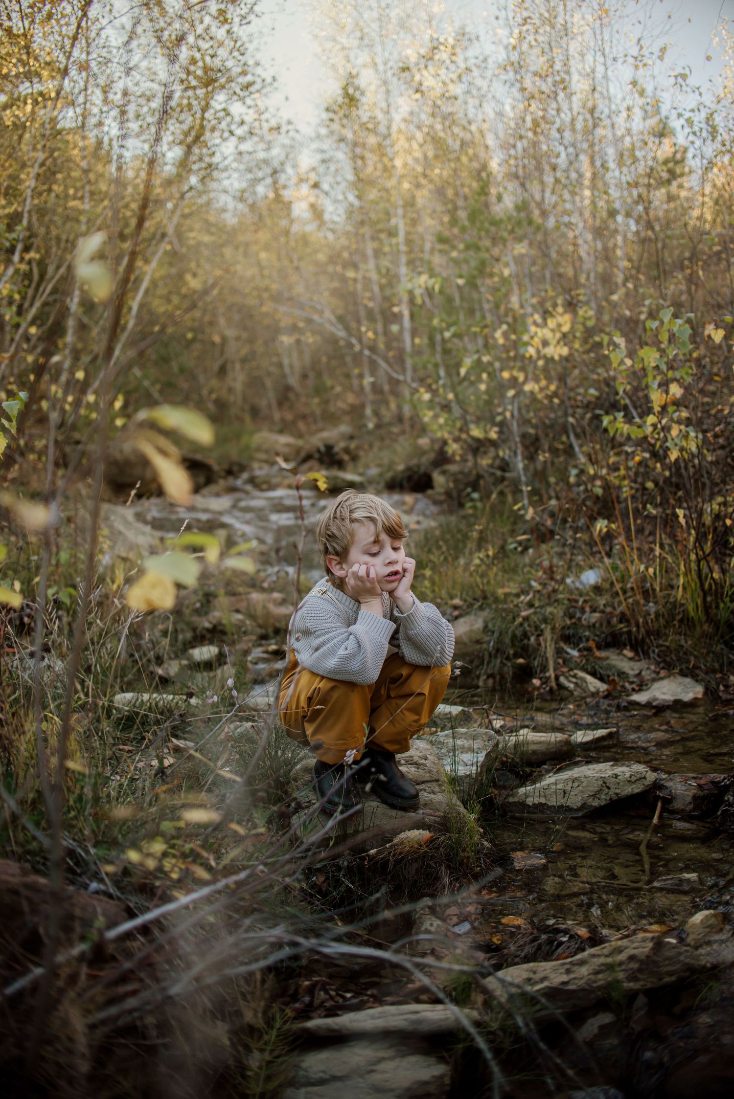 Child in mustard overalls crouching among rocks by forest stream in autumn setting.