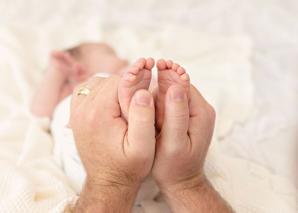 Adult hands gently holding tiny baby feet against soft white fabric background.