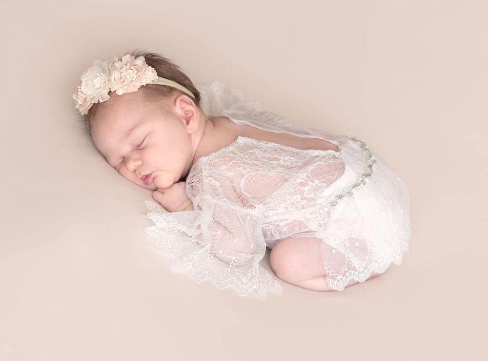 Sleeping newborn wearing delicate white lace romper and headband rests peacefully on soft pink backdrop.