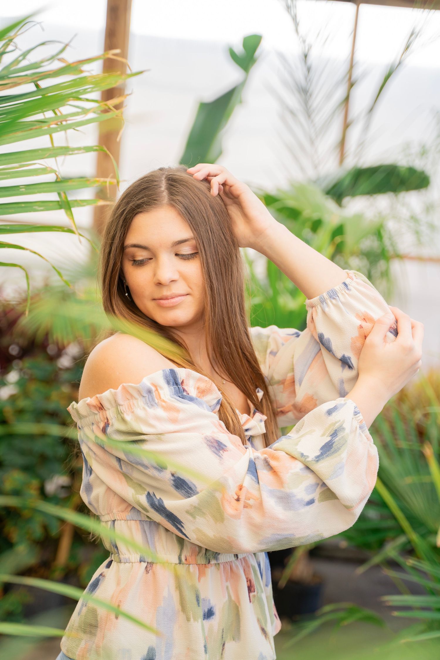 Brunette woman in floral dress posing among tropical plants in a greenhouse setting.