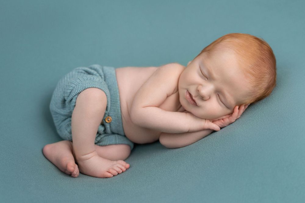 Peaceful sleeping baby in blue shorts posed on teal backdrop.