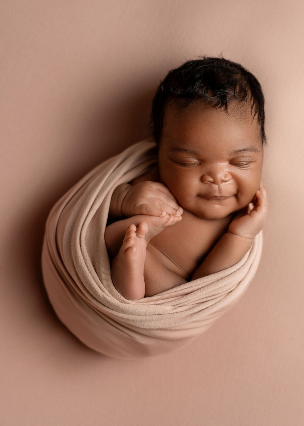 Sleeping newborn wrapped in cream fabric during artistic portrait session on pink background.