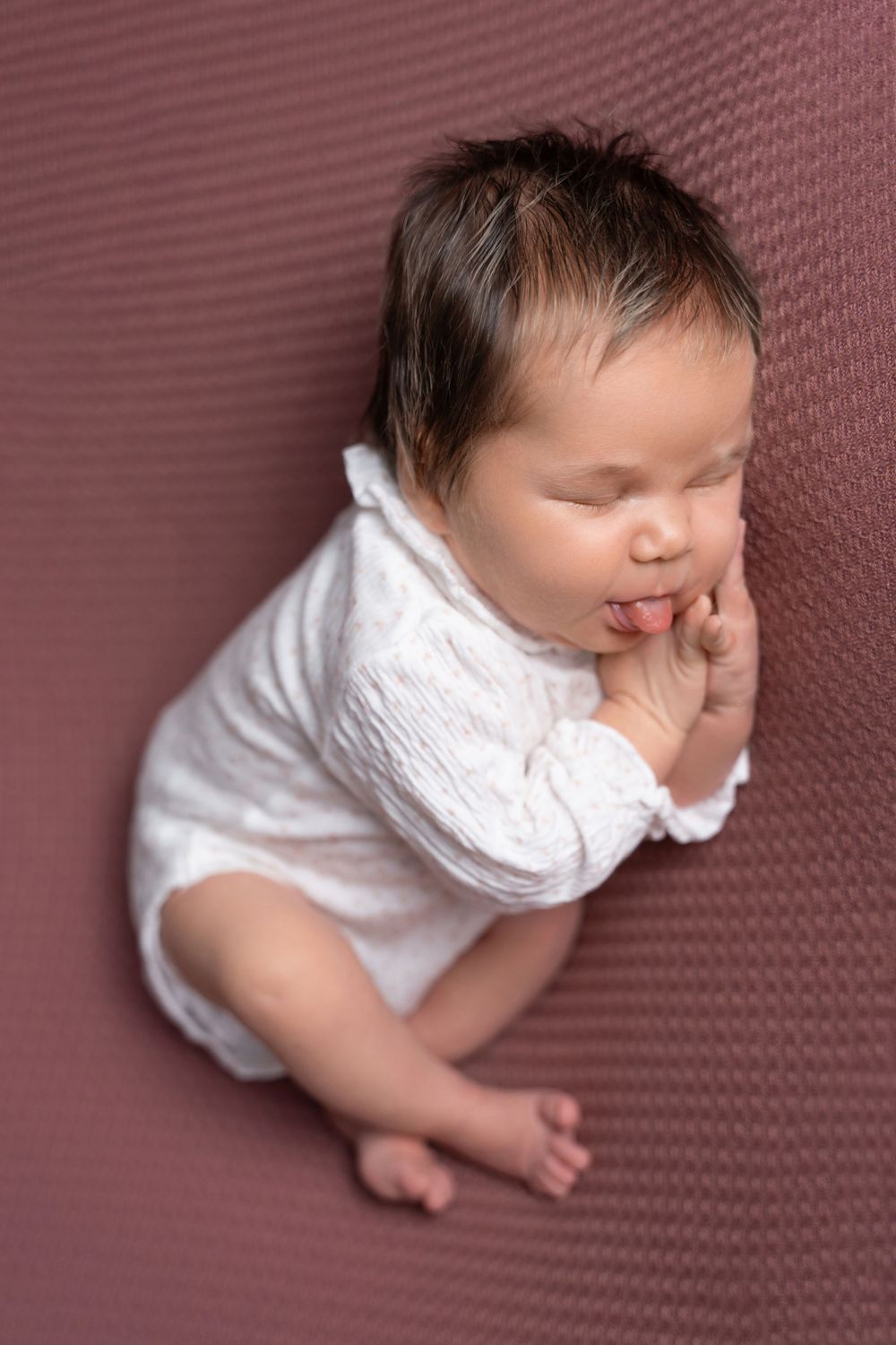 Baby in white outfit lying on burgundy textured backdrop.