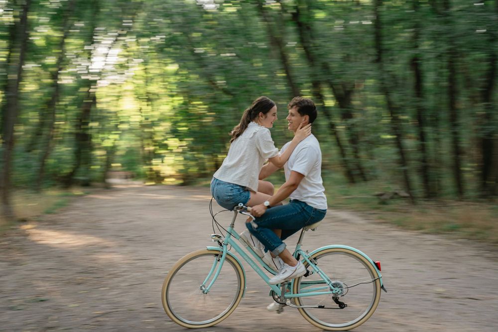 Two people share a vintage mint-colored bicycle while riding together along a tree-lined forest path.