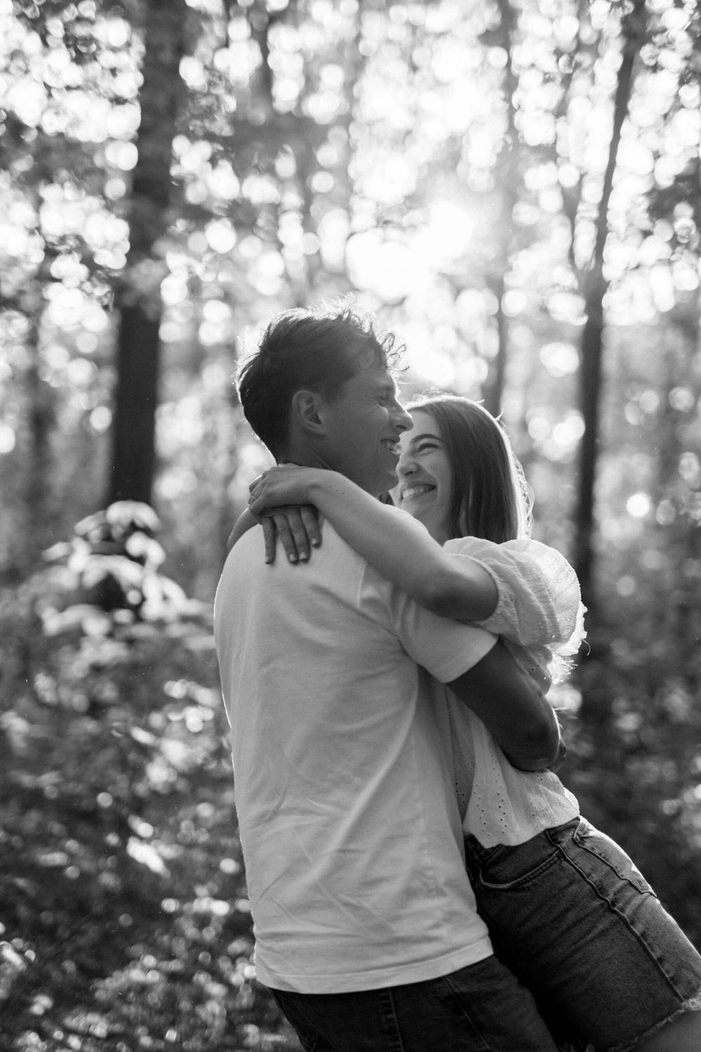 Black and white intimate portrait of couple embracing in forest with bokeh light effects.