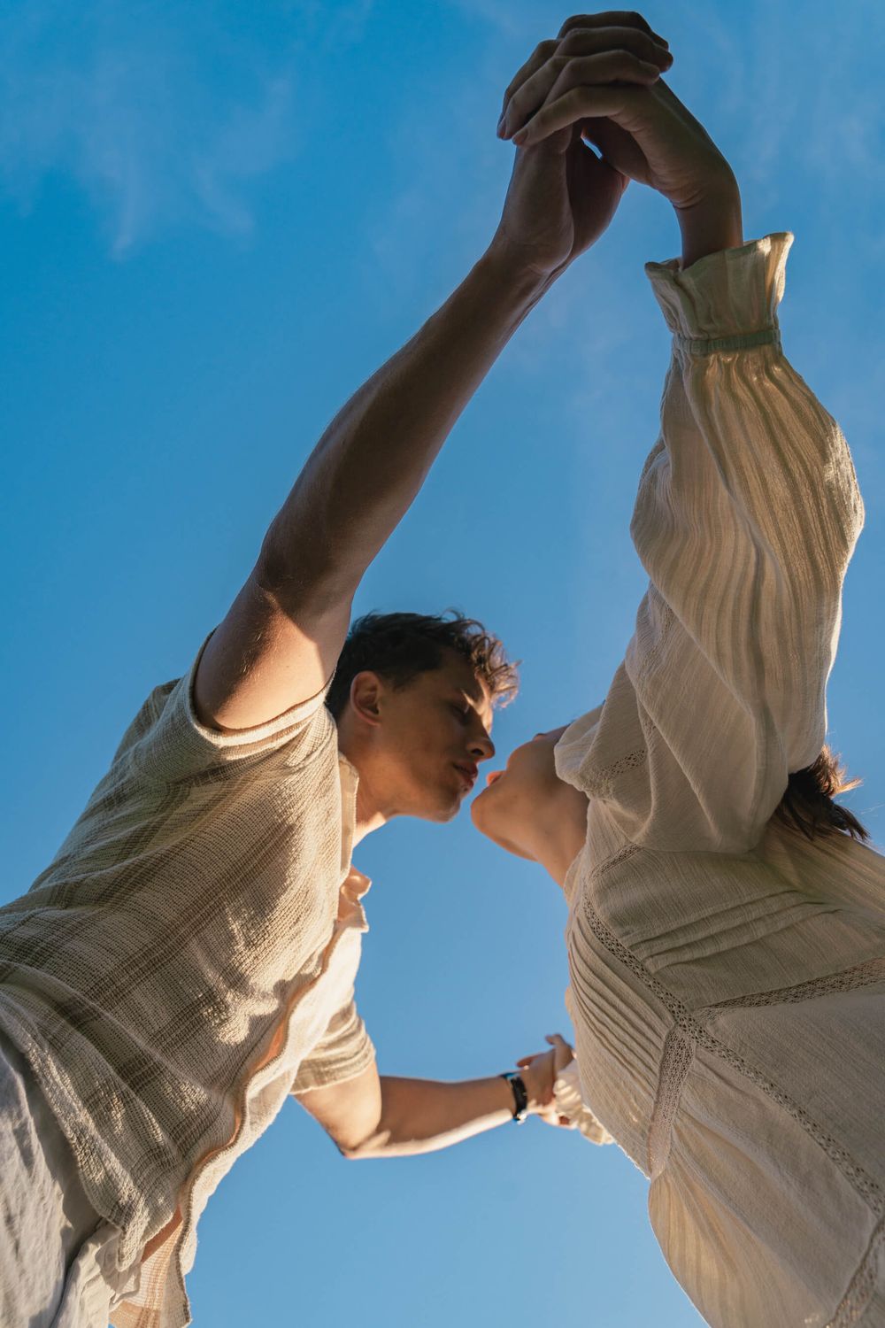 Two people in white clothing reach their arms up to the sky against a bright blue background.