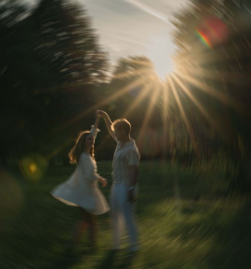 Couple dancing in a meadow at sunset with sunburst rays shining through trees creating a dreamy atmosphere.