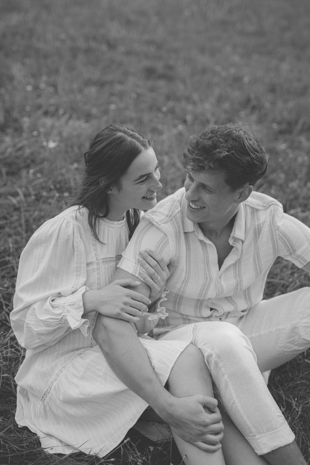 Couple in white clothing share a tender moment while sitting in a grassy field.