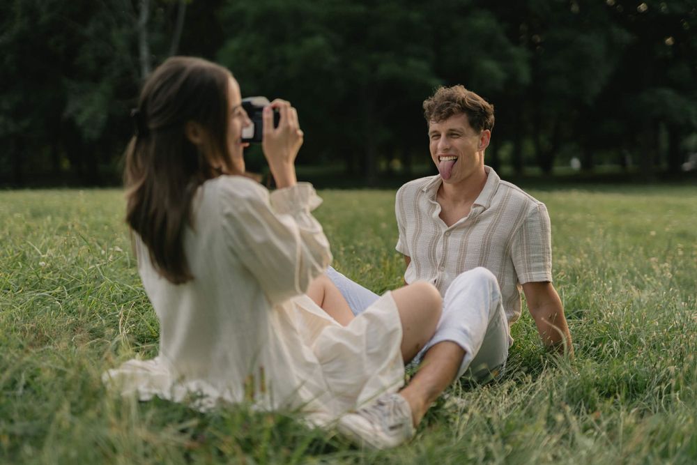 Two people in white clothing taking photos of each other while relaxing on grass.