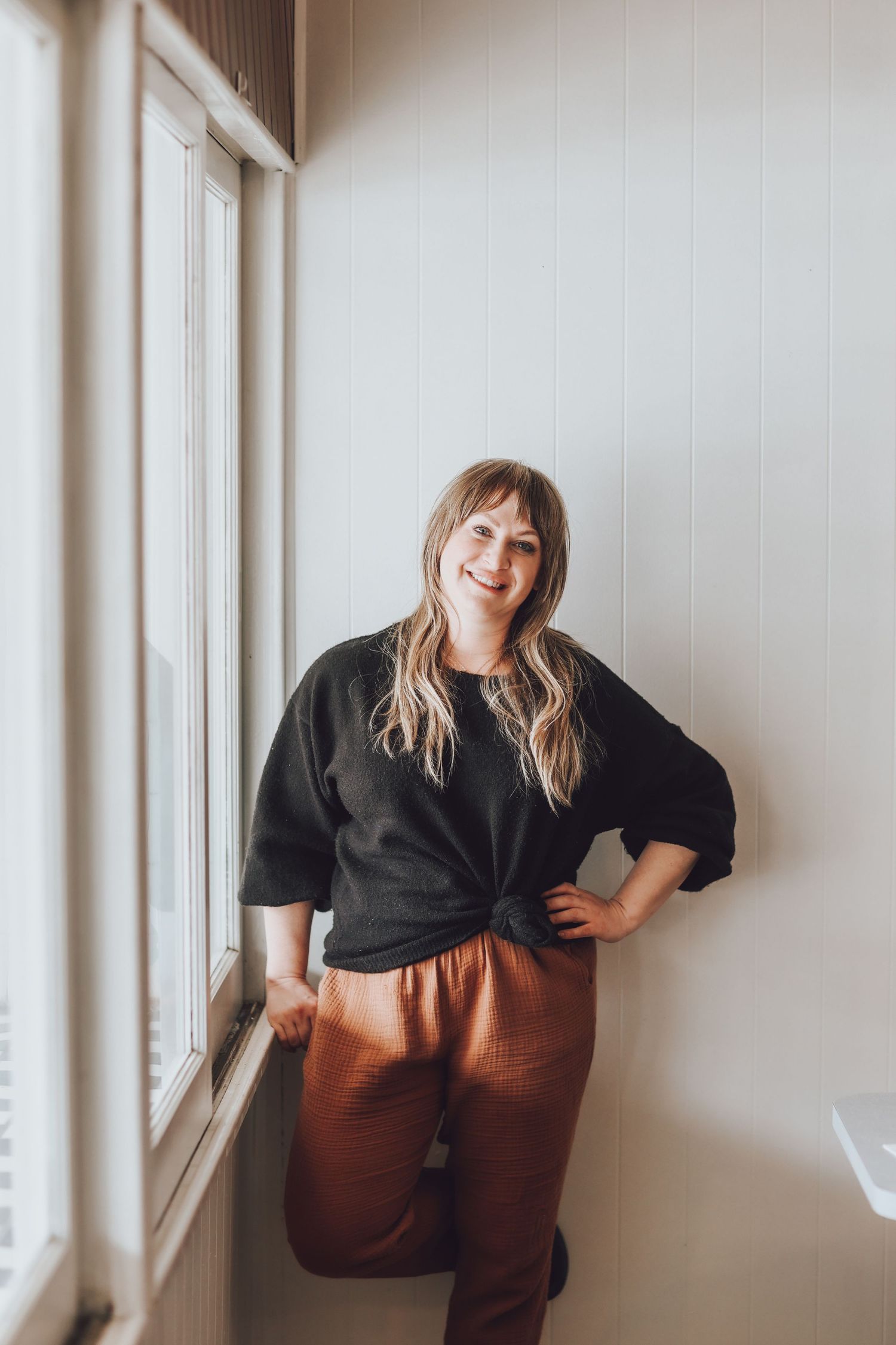 Candid photo shoot against white wall showing person in black top and brown pants in multiple poses.