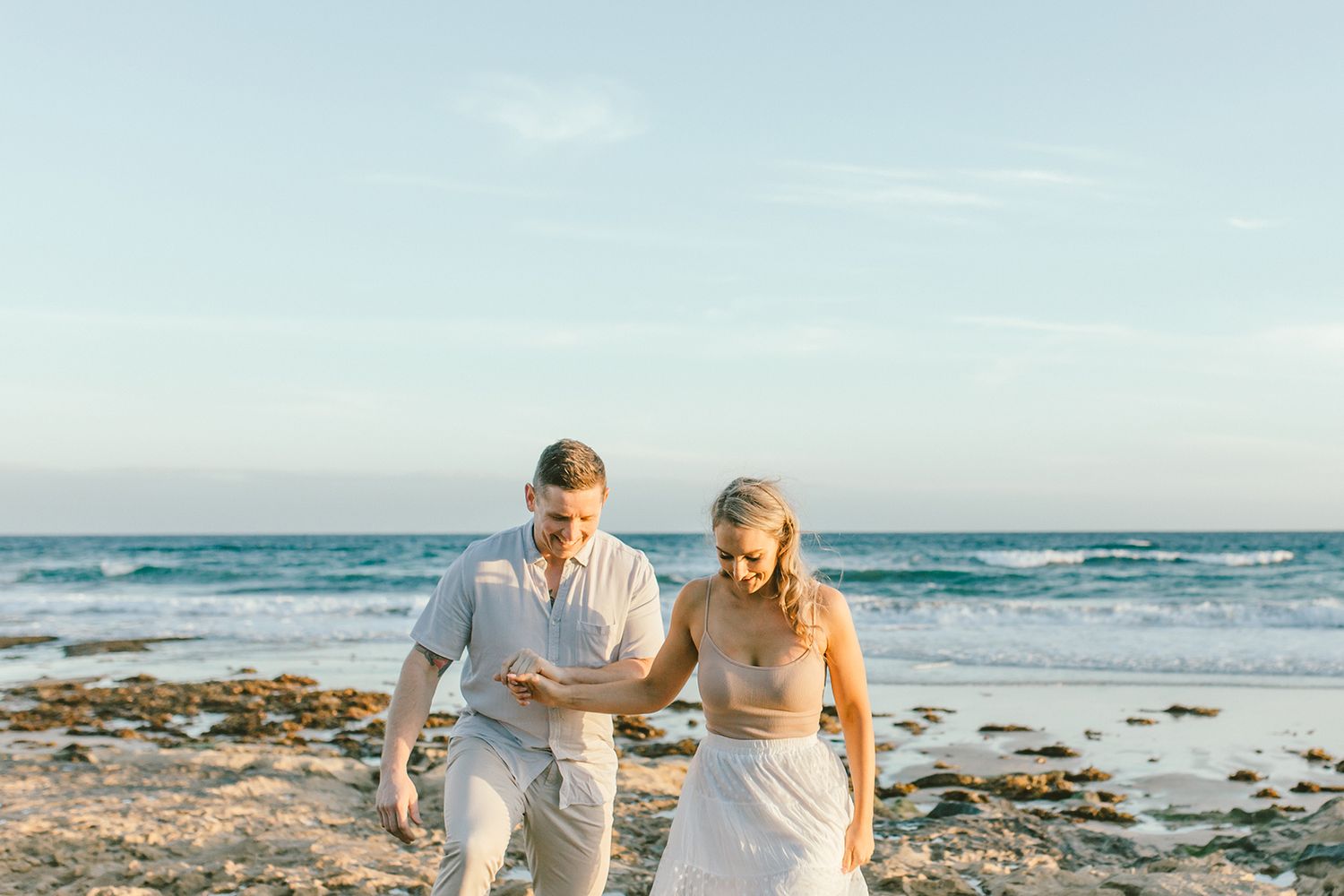 A couple strolls together on a rocky beach with turquoise ocean waves in the background.