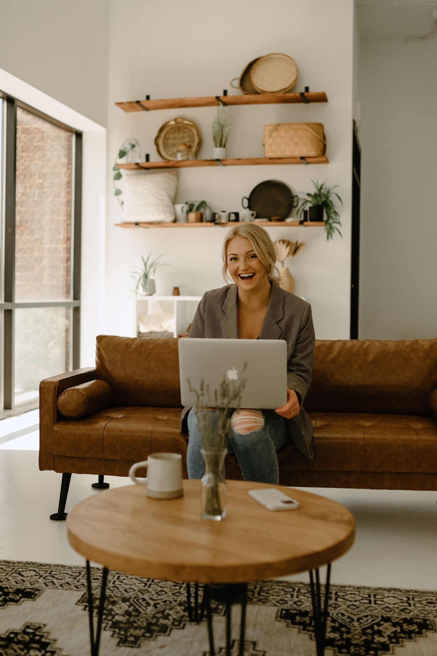 Woman working on laptop while sitting on brown leather couch with decorative wall shelves behind her.