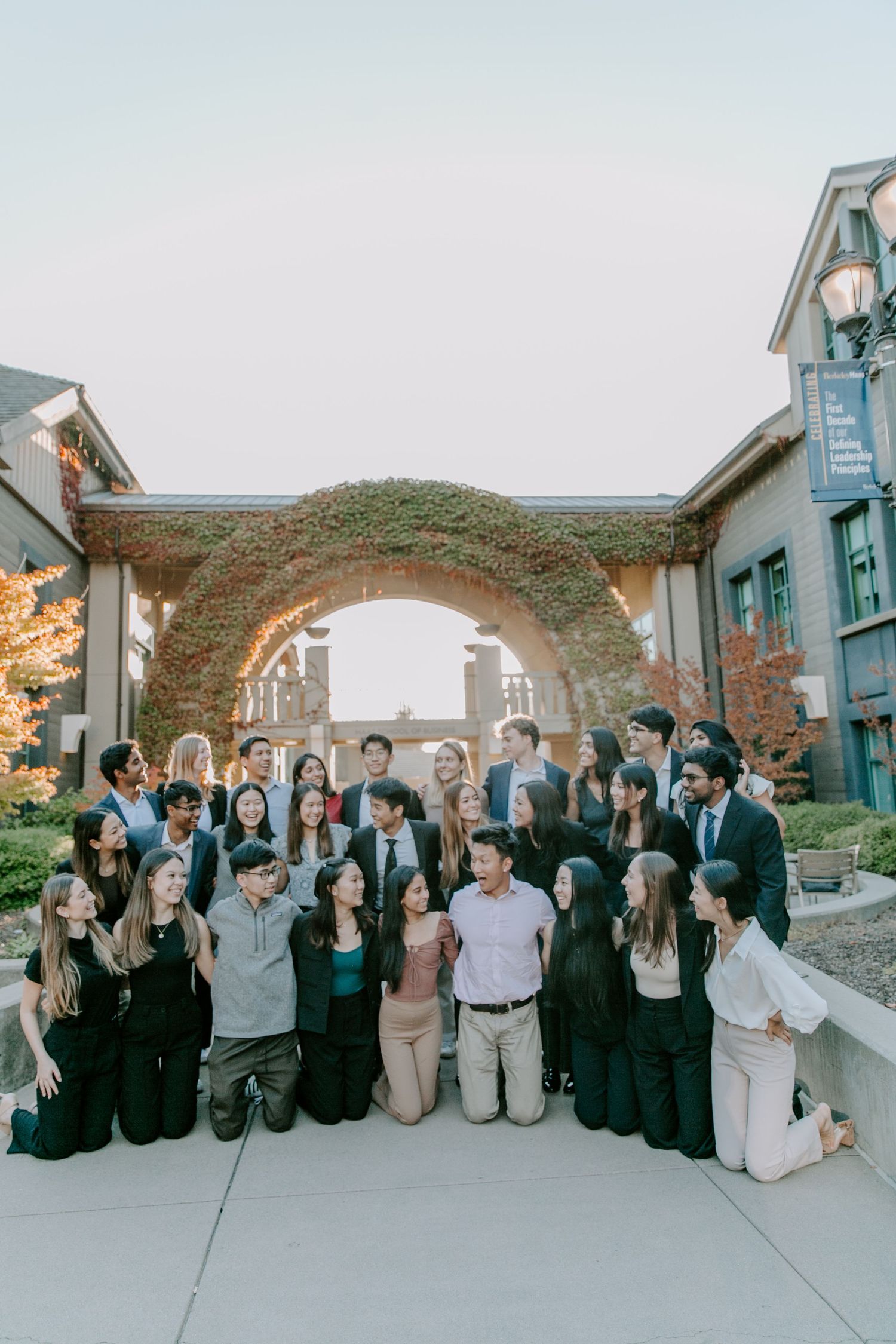 Large group poses together in front of an ivy-covered stone archway on a college campus.