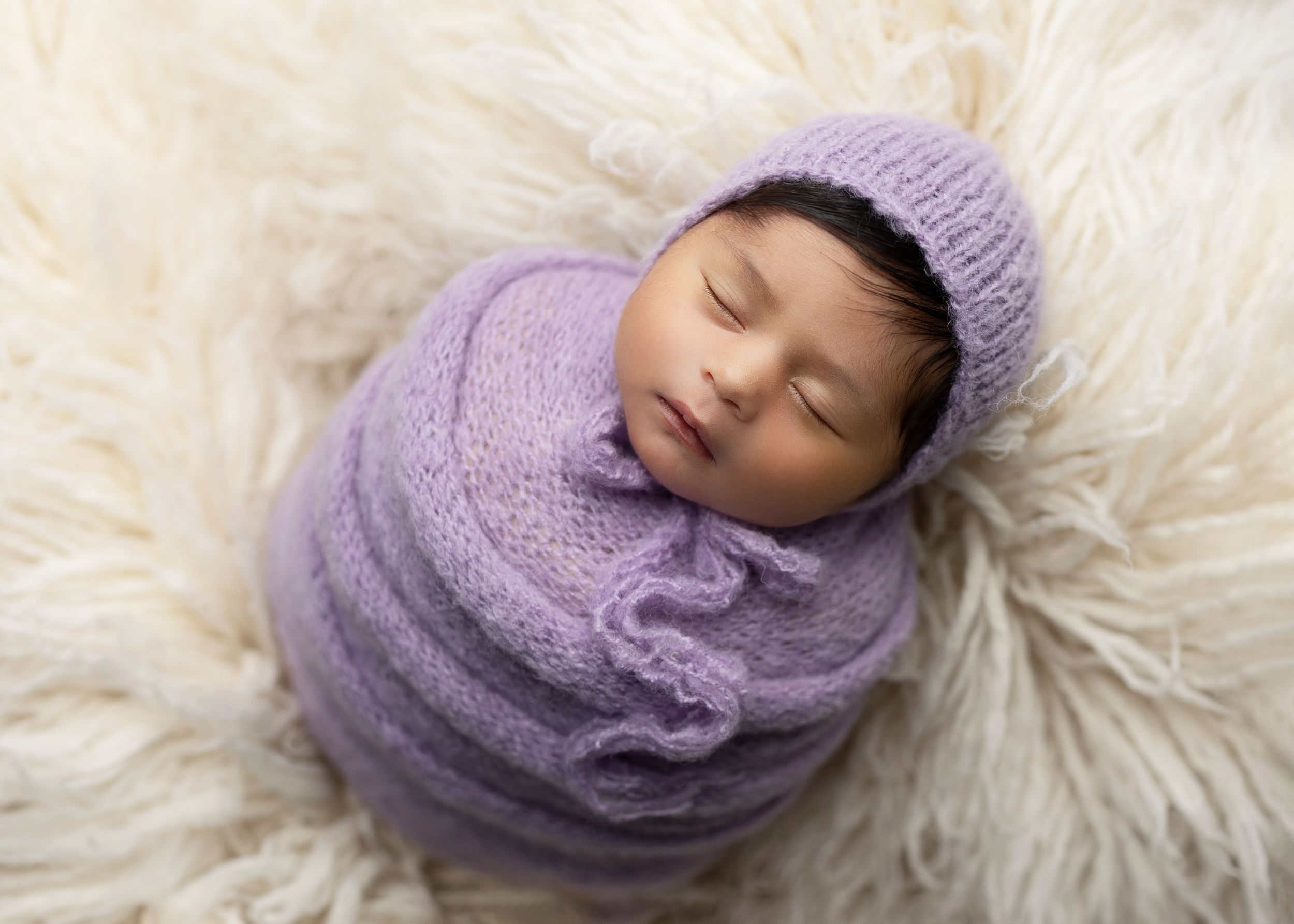 Newborn baby wearing purple knit hat and wrap on soft white fur.