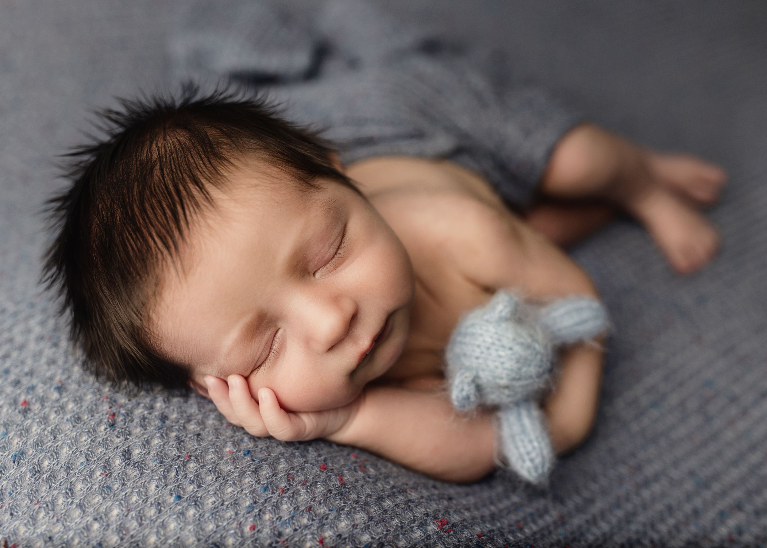 A sleeping newborn baby rests on a gray blanket with a small knit accessory.