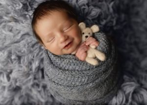 A smiling newborn swaddled in gray knit wrap with tiny plush toy.