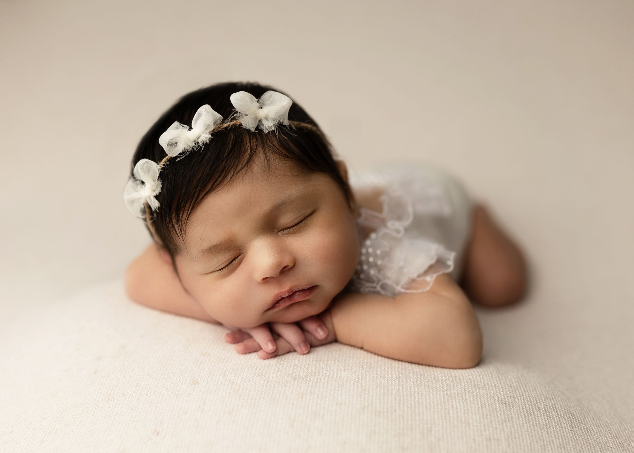 A sleeping newborn baby wearing a white floral headband rests peacefully on cream-colored fabric.