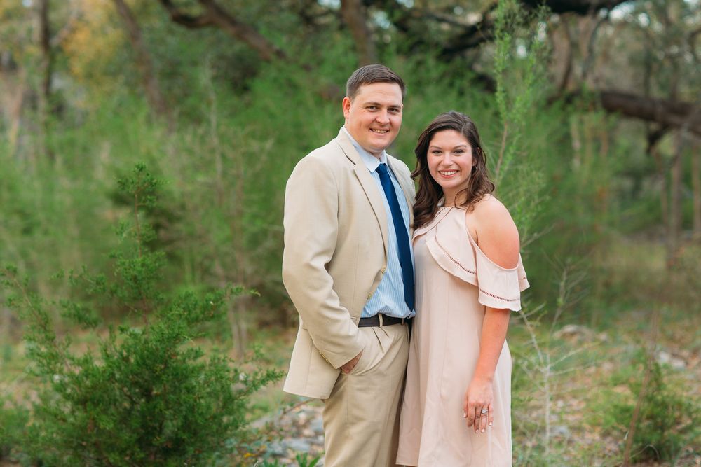 A couple poses together outdoors in formal attire with lush green trees and foliage in the background.