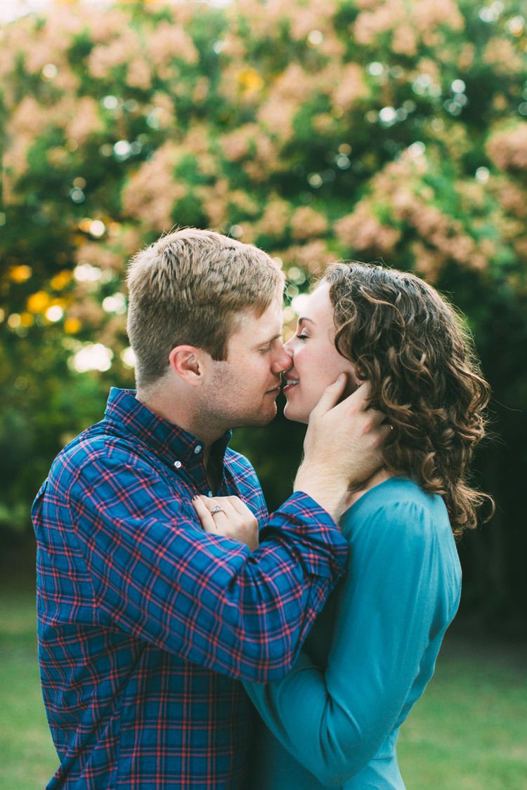 A couple wearing a blue plaid shirt and turquoise sweater share a romantic kiss outdoors in autumn.