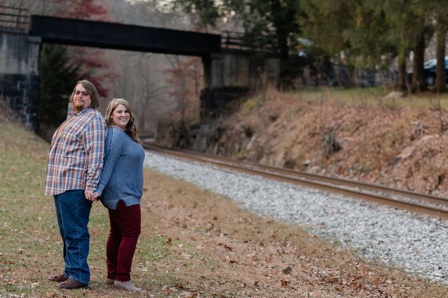 A couple stands together near railroad tracks with an industrial bridge in the background.