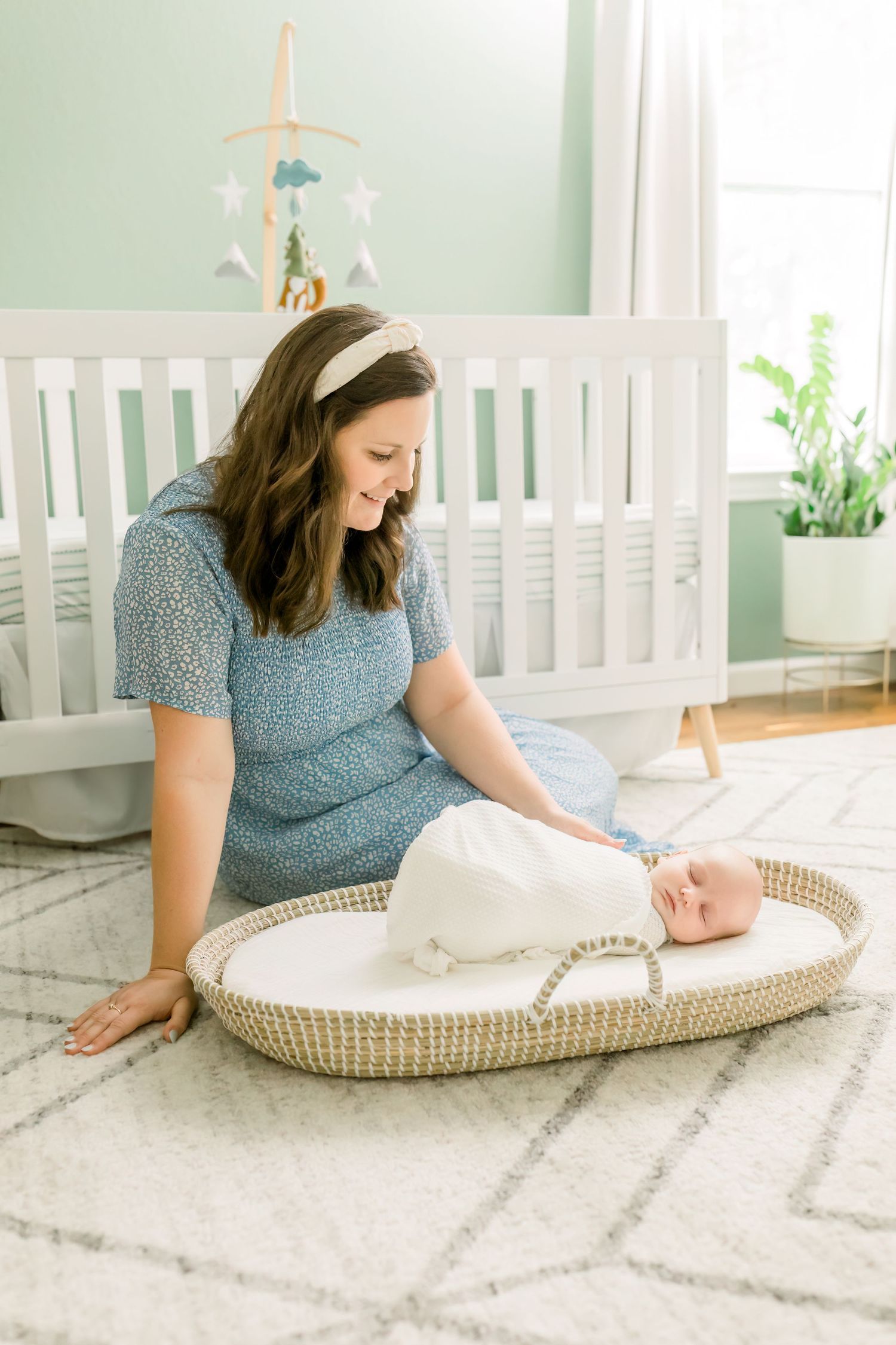 Nursery scene with white crib, mint walls, and woman in blue dress sitting beside baby basket.