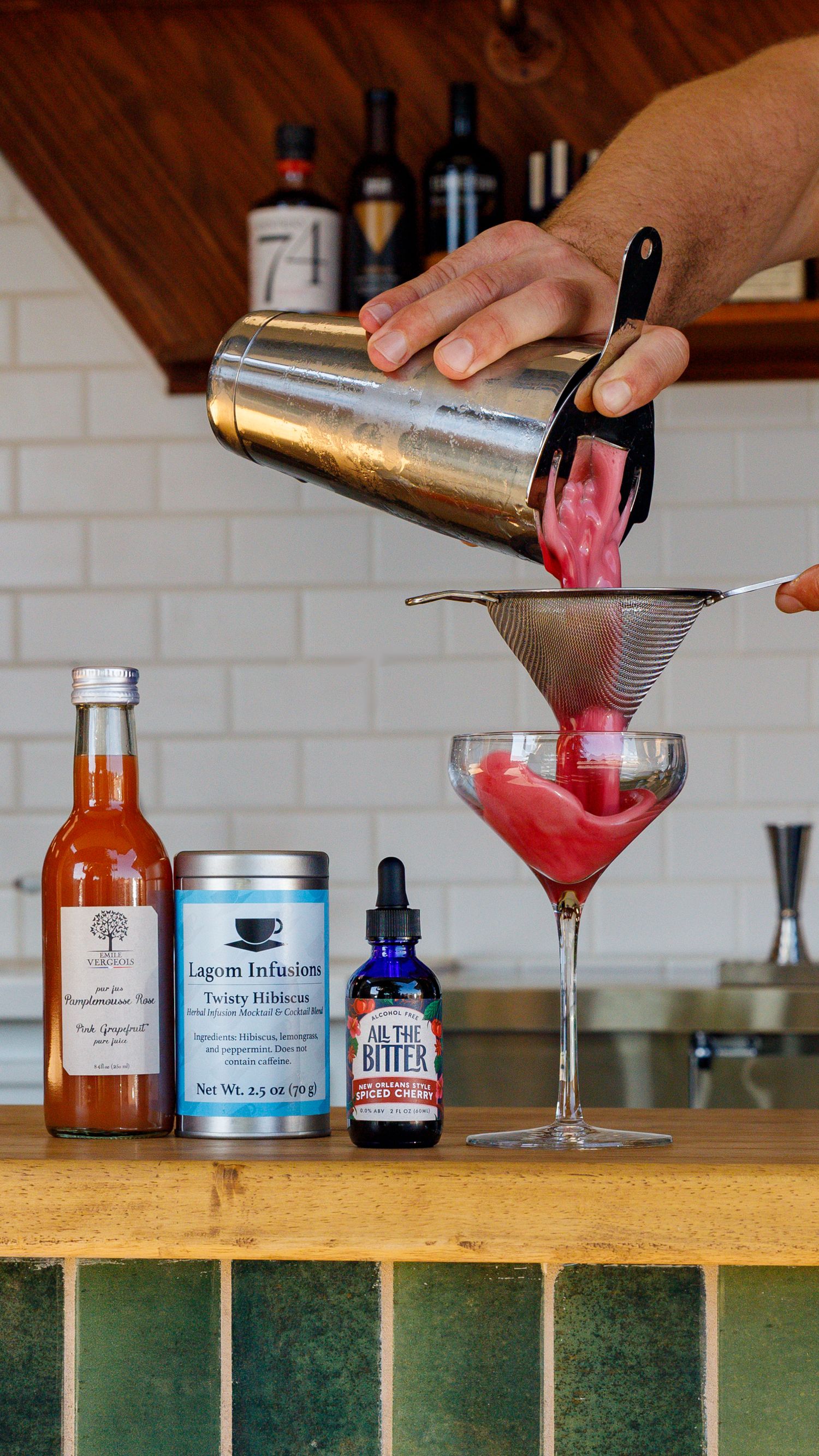 A pink cocktail being strained through a fine mesh strainer into a martini glass with bottles and mixers in background.