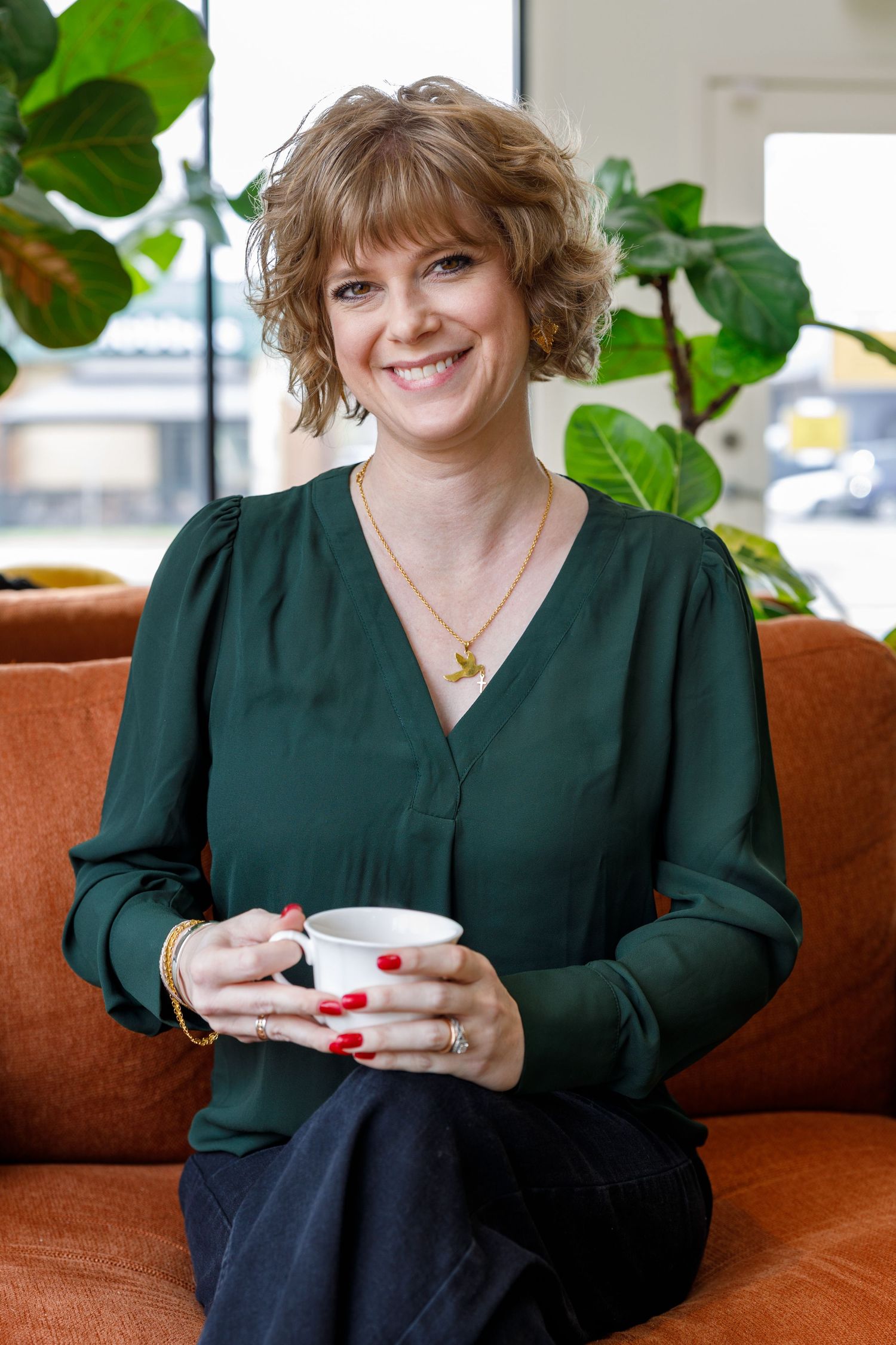 A person in a dark green blouse sits on an orange couch holding a white coffee cup, with houseplants in the background.