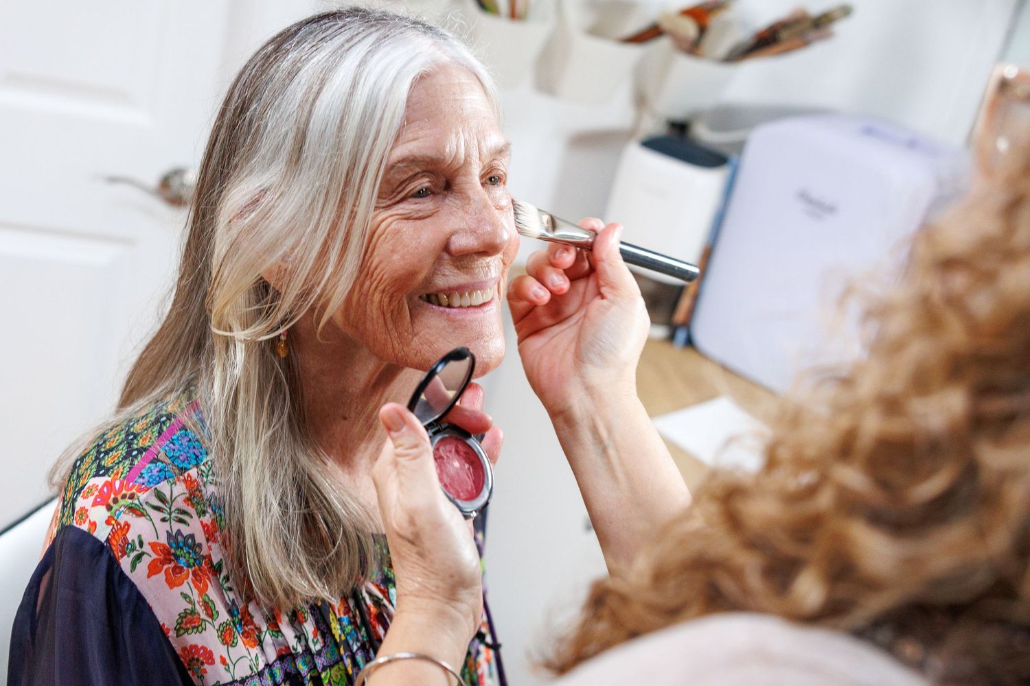 Professional makeup artist applying cosmetics to mature woman with silver hair in floral dress during beauty session.