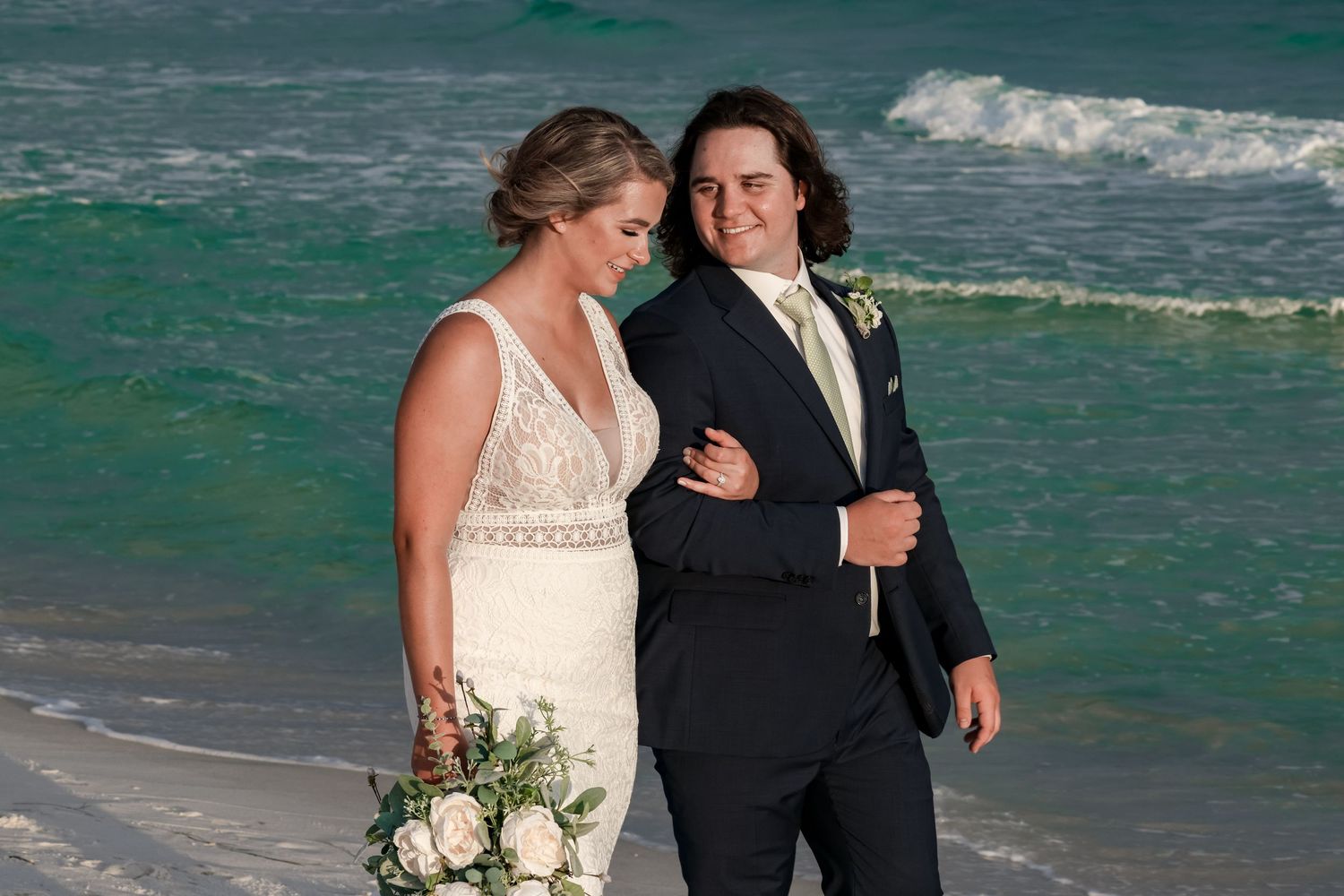 Newlyweds share an intimate moment together on a beach with turquoise waves crashing behind them.