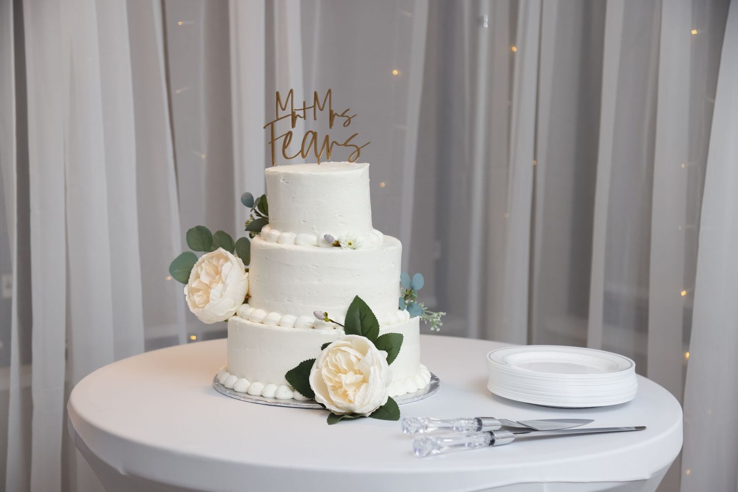 Elegant white wedding cake decorated with roses and a 'Mr & Mrs' topper on a round table.